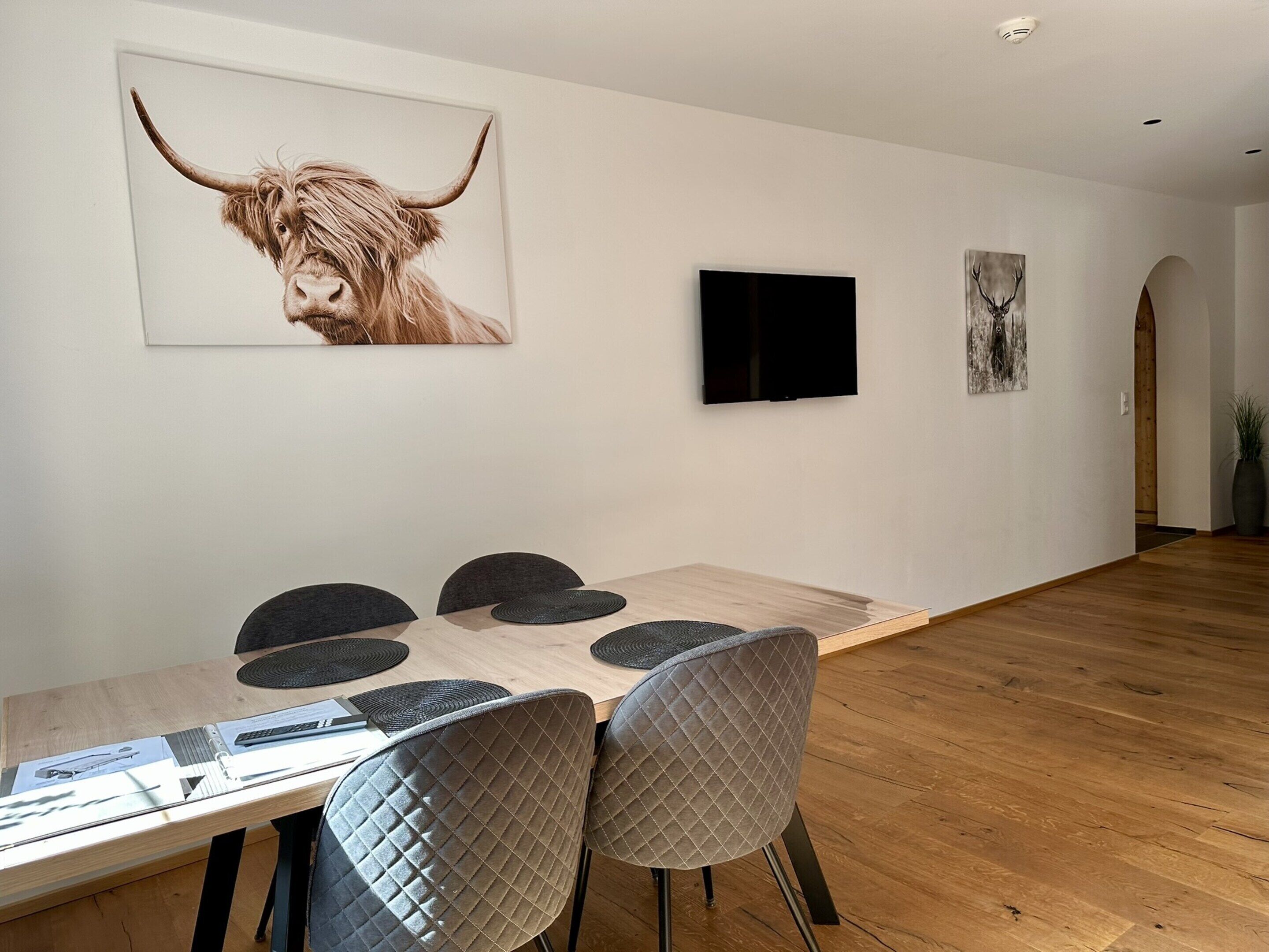 A dining area with a wooden table, quilted chairs, artwork on the wall, and a flat-screen TV.