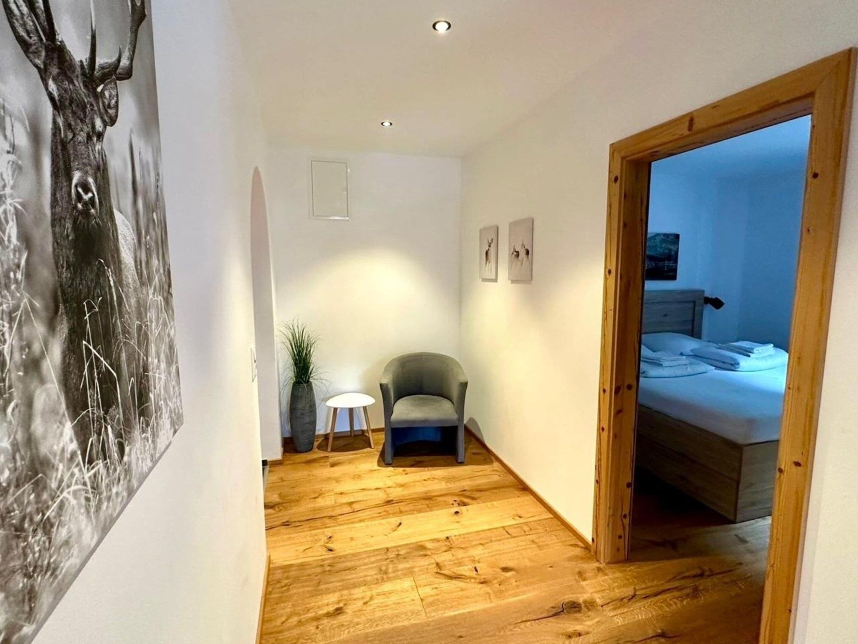 A narrow hallway with a wooden floor, a grey armchair, a small table with a lamp, and a bed in the adjacent room.