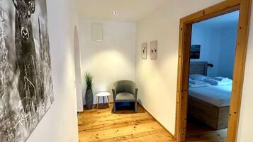 A narrow hallway with a wooden floor, a grey armchair, a small table with a lamp, and a bed in the adjacent room.