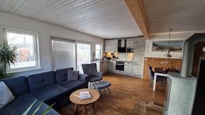 A modern living room with a sofa, coffee table, and a dining area with chairs. There is a kitchen with appliances and a large window. - Holiday Home Apartment - East (Grossenbrode)