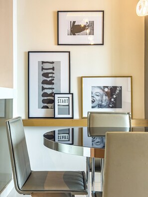 Een moderne eetruimte met een glazen tafel, metalen stoelen en kunstwerken in lijsten aan de muur.