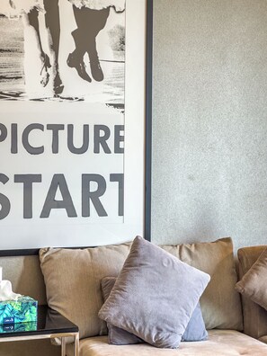 Een lijst met een kunstwerk waarop de tekst 'PICTURE START' staat, een beige bank met meerdere kussens en een kleine tafel met een zakdoekendoos.