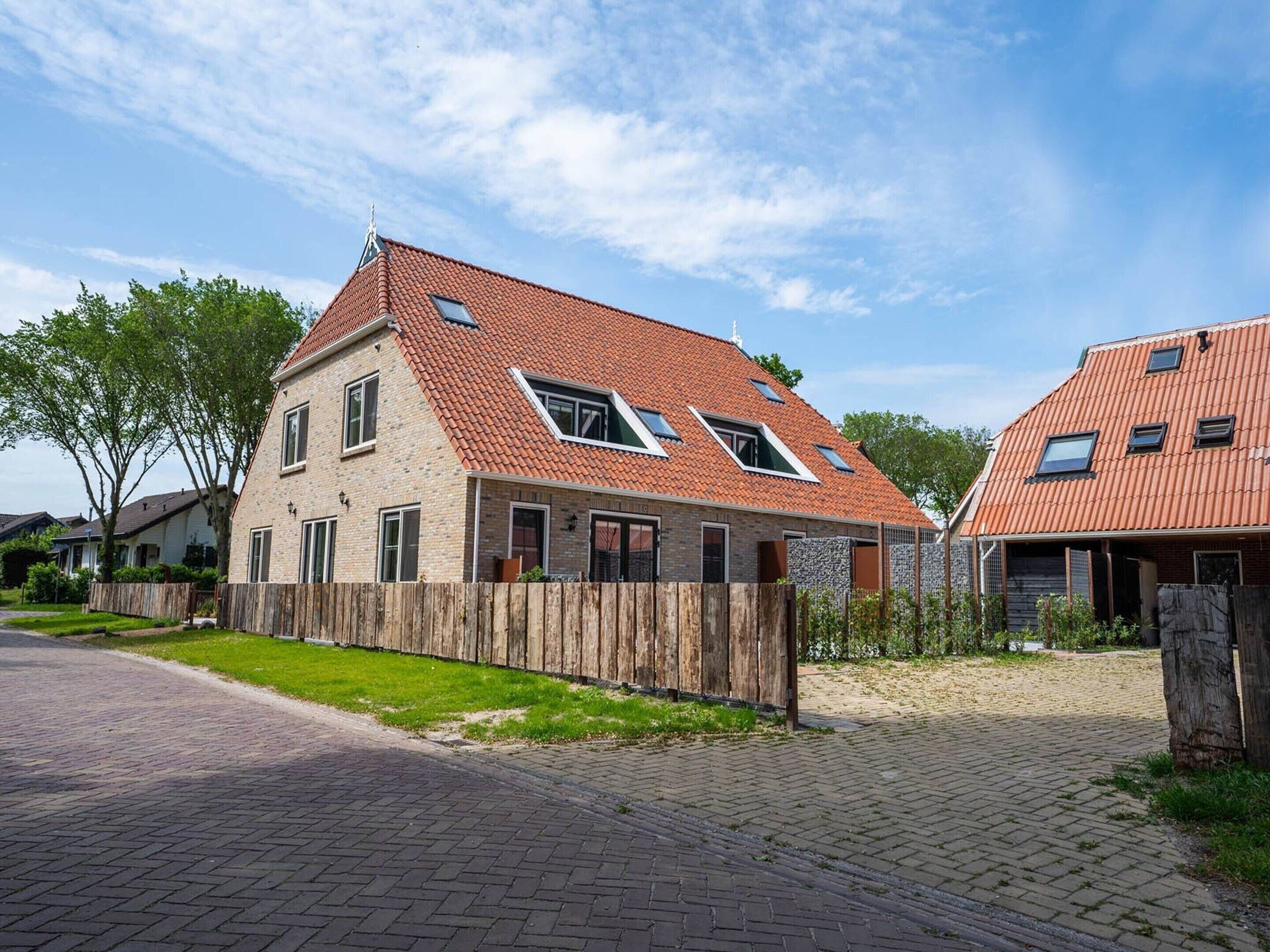 Een tweelaags huis met een rode pannendak, een houten hek en een verharde oprit.