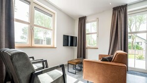 Un salon moderne avec un canapé, un fauteuil et une table basse. De grandes fenêtres sont présentes, ornées de rideaux, et un téléviseur à écran plat est fixé au mur. On peut admirer la vue sur l’extérieur.