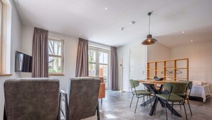 Un espace de repas moderne avec une table et des chaises en bois, un téléviseur fixé au mur et de grandes fenêtres avec des rideaux.