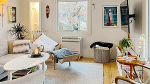 Ein gemütliches Wohnzimmer mit einer Couch, einem kleinen Tisch mit einer Kerze, einem Korbstuhl und Blick ins Freie durch ein Fenster.