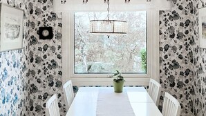 Ein Essbereich mit einem weißen Tisch, gestreiften Stühlen, einer Topfpflanze, einer Uhr und einem Fenster mit Blick auf Bäume.