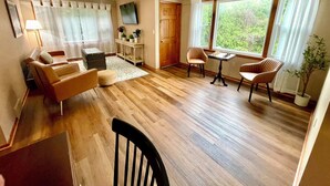 Een ruime woonkamer met houten vloer, een bank, een fauteuil, een klein tafeltje met een schaakspel en een aan de muur bevestigde televisie.