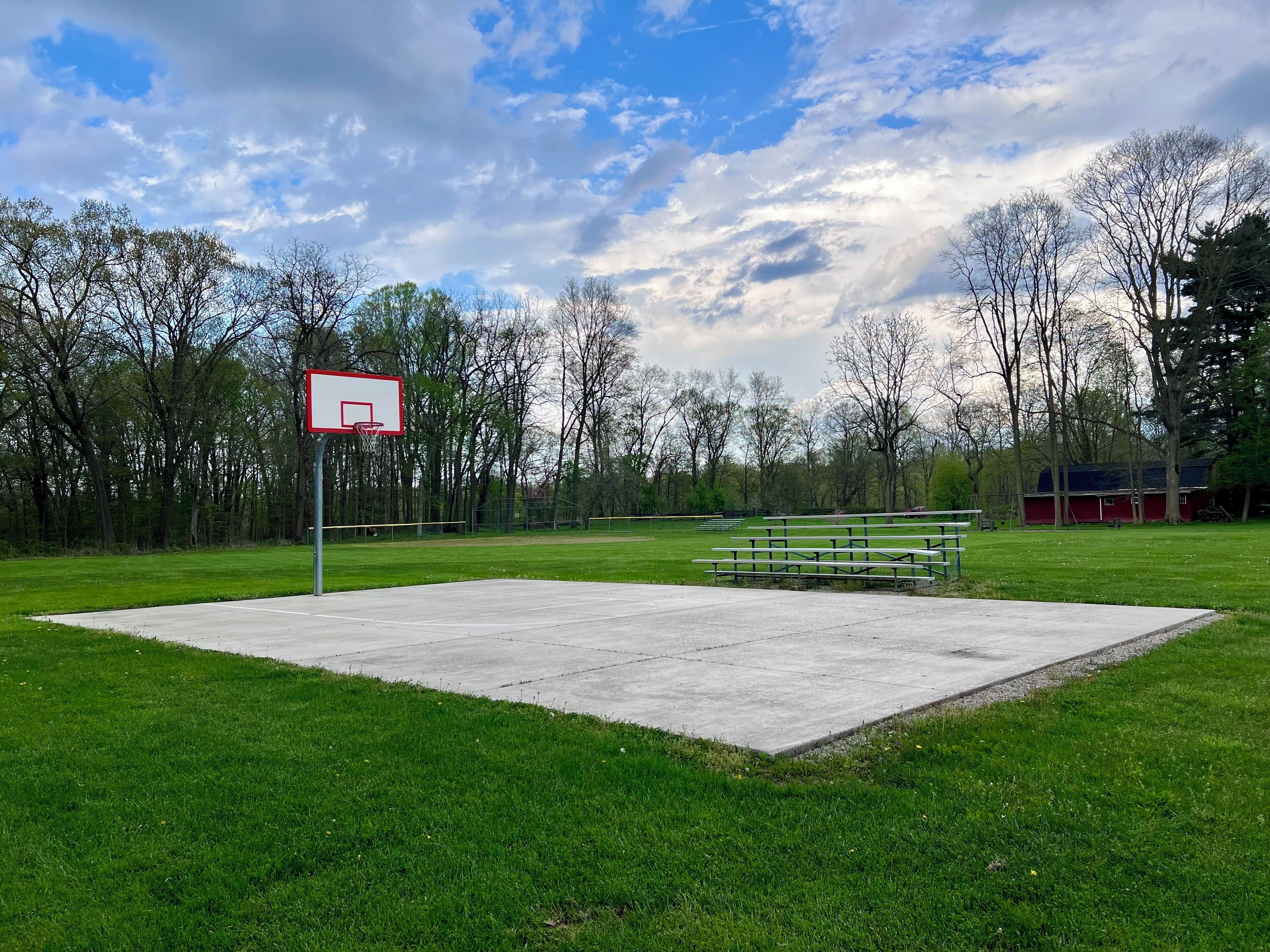 A basketball court with a hoop, benches, and a grassy area with trees in the background.