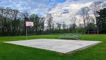 A basketball court with a hoop, benches, and a grassy area with trees in the background.