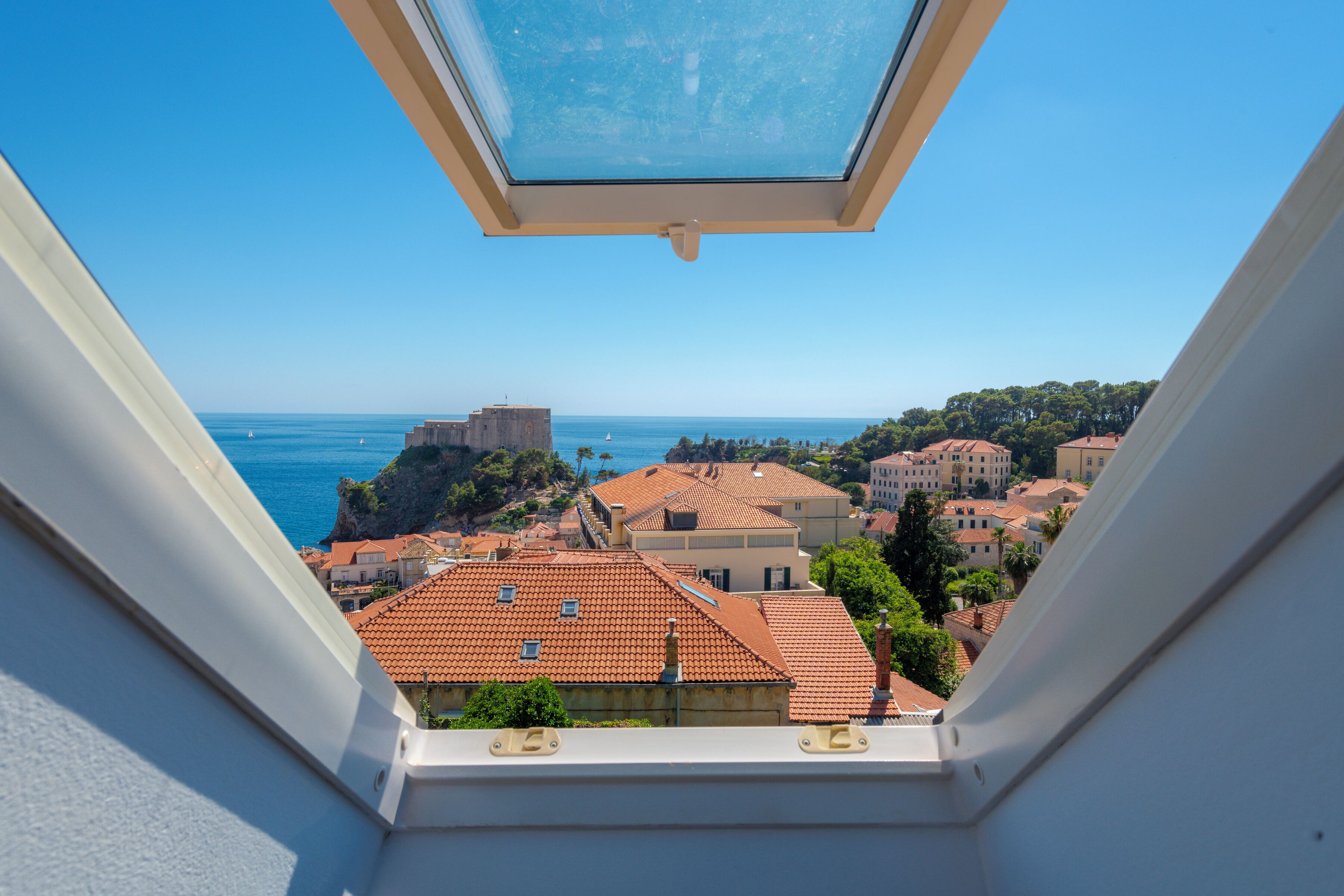 A view from a window overlooking a coastal town with terracotta-roofed buildings and a clear blue sky.