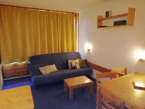 Un salon avec un canapé bleu foncé, deux chaises en bois, une petite table en bois et un rideau jaune.