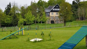 A grassy outdoor area with a blue slide and yellow and blue poles, a stone building with a black roof in the background, and trees around. - Mrs. Monika Rasehorn (Altenberg)