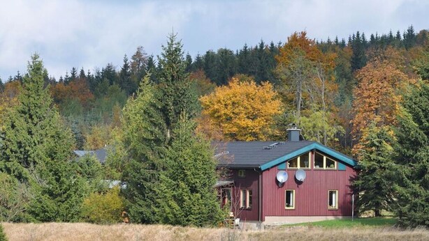 A single-story house with a dark roof and a red exterior, surrounded by dense forest and autumn foliage. - Mrs. Monika Rasehorn (Altenberg)