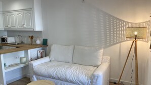 Ein weißes Sofa, ein kleiner runder Holztisch und eine Stehlampe in einem Zimmer mit Küchenbereich.