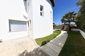 A white building with a garage door, a paved driveway, and a well-maintained lawn with a palm tree. - Nice Apartment with Pool (Siracusa)