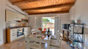 Keittiössä on puupalkit katossa, valkoiset kaapit, ruokapöytä tuoleineen ja avoimesta ovesta avautuva näkymä ulos.