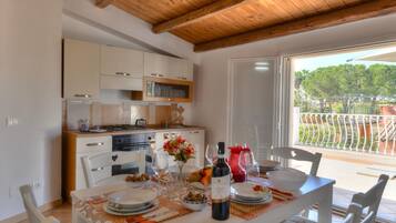 A dining area with a table set for a meal, a kitchen in the background, and a view of a balcony with a railing.