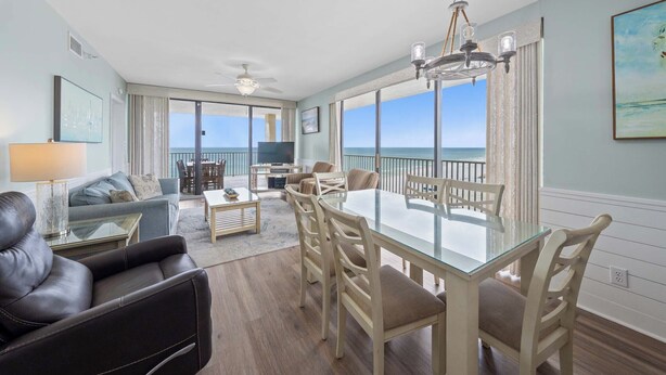 A spacious living area with a glass dining table, comfortable seating, a TV, and a balcony with a view of the ocean. - 🌊 Romar Place 1106 | Top Floor Gulf Front | Corner Balcony + Resort Amenities (Orange Beach)