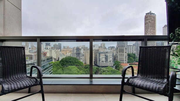 A balcony with two wicker chairs and a cityscape view through a large window. - Vossa Bossa Setin República (São Paulo)