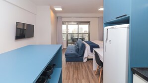 A compact hotel room with a blue sofa, a small dining table, a flat-screen TV, a refrigerator, and a view of the city through large windows. - Vossa Bossa Setin República (São Paulo)