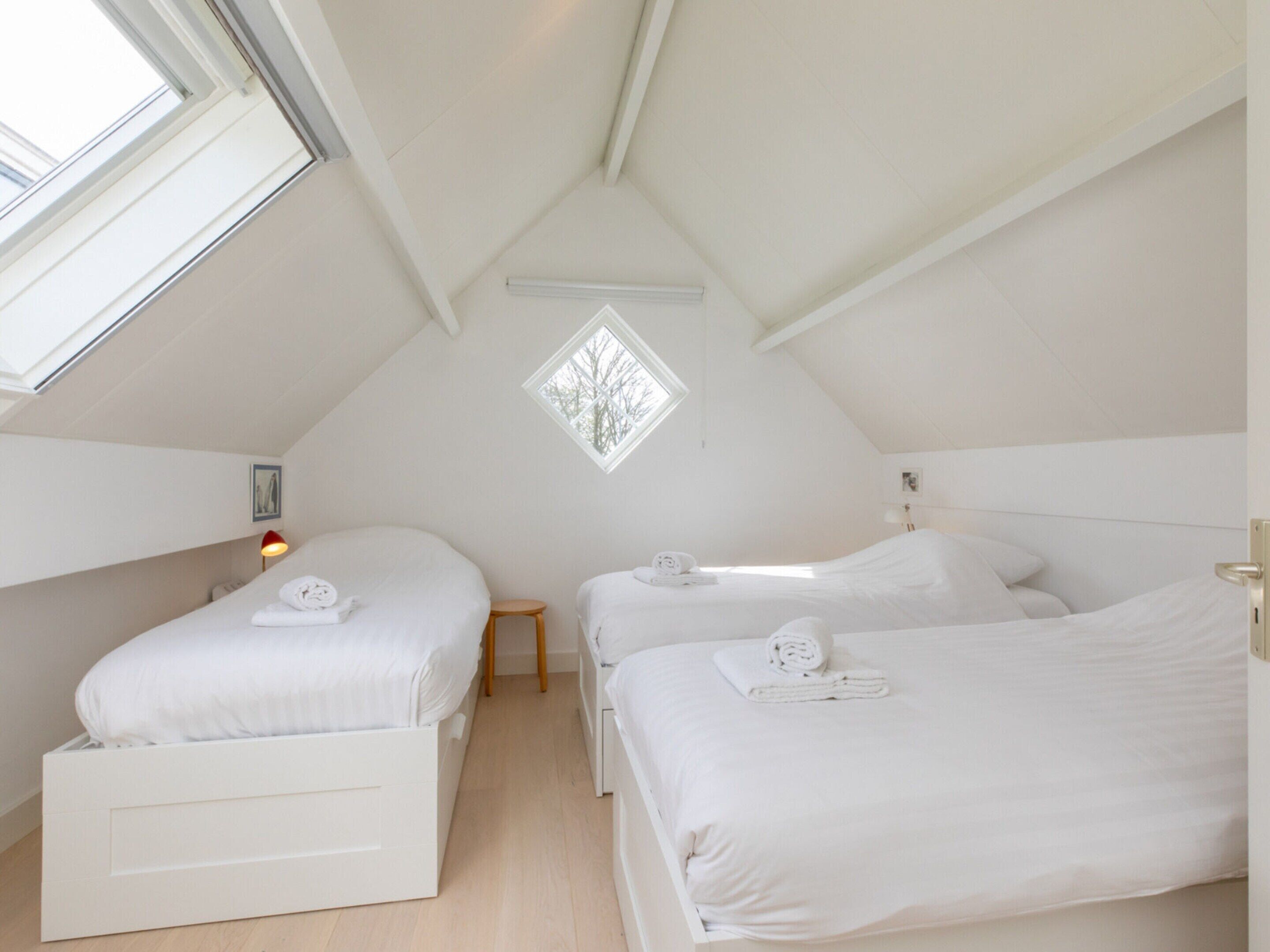 Une petite chambre mansardée, aux murs blancs, avec deux lits, un puits de lumière et une petite fenêtre.