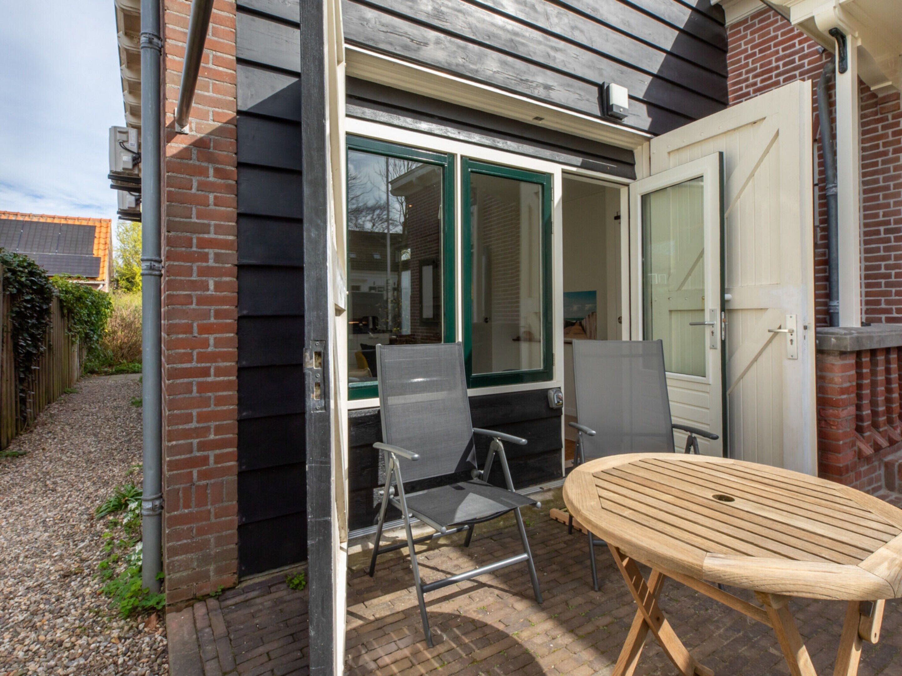 Une terrasse avec deux chaises et une table, une porte et une fenêtre avec des stores.