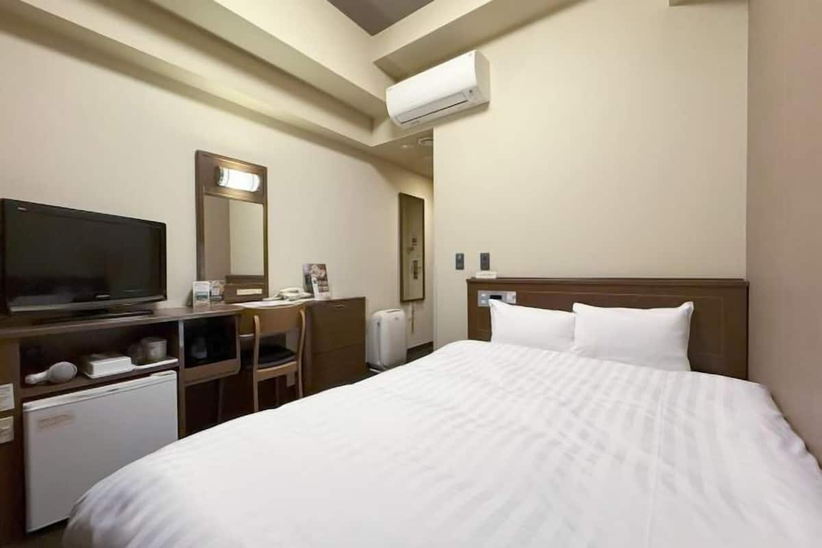 Ein Hotelzimmer mit Bett, Fernseher, Schreibtisch, Stuhl, kleinem Kühlschrank und Klimaanlage.