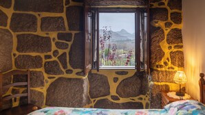 A room with a stone wall, a wooden bed with a floral bedspread, a window with a view of mountains, and a bedside lamp. - GerêsTER  (Terras de Bouro)