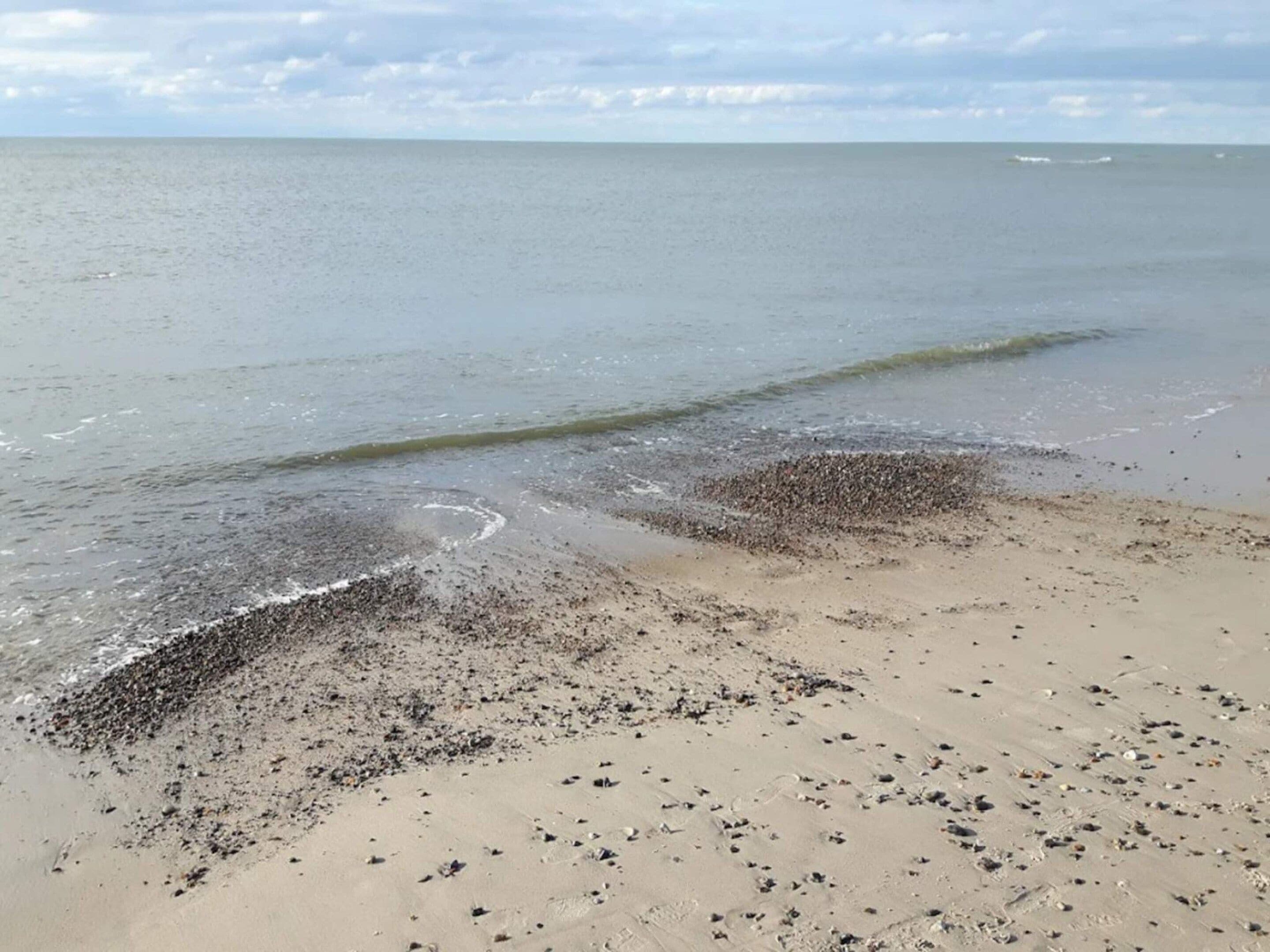 A beach with pebbles and sand, calm water, and a cloudy sky.