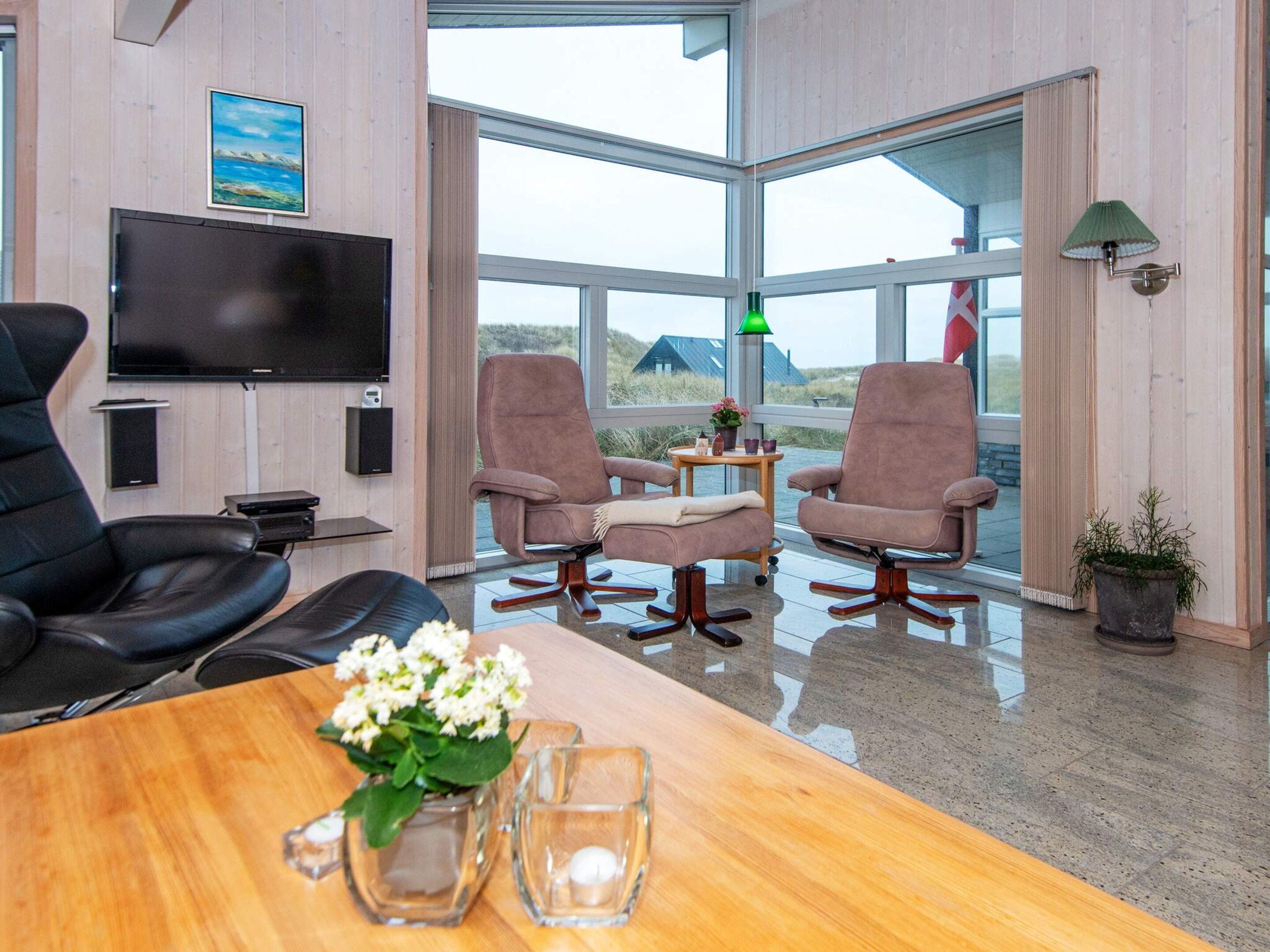 A modern room with a large TV, two leather chairs, a wooden table with a vase of flowers, and a view of the outdoors.