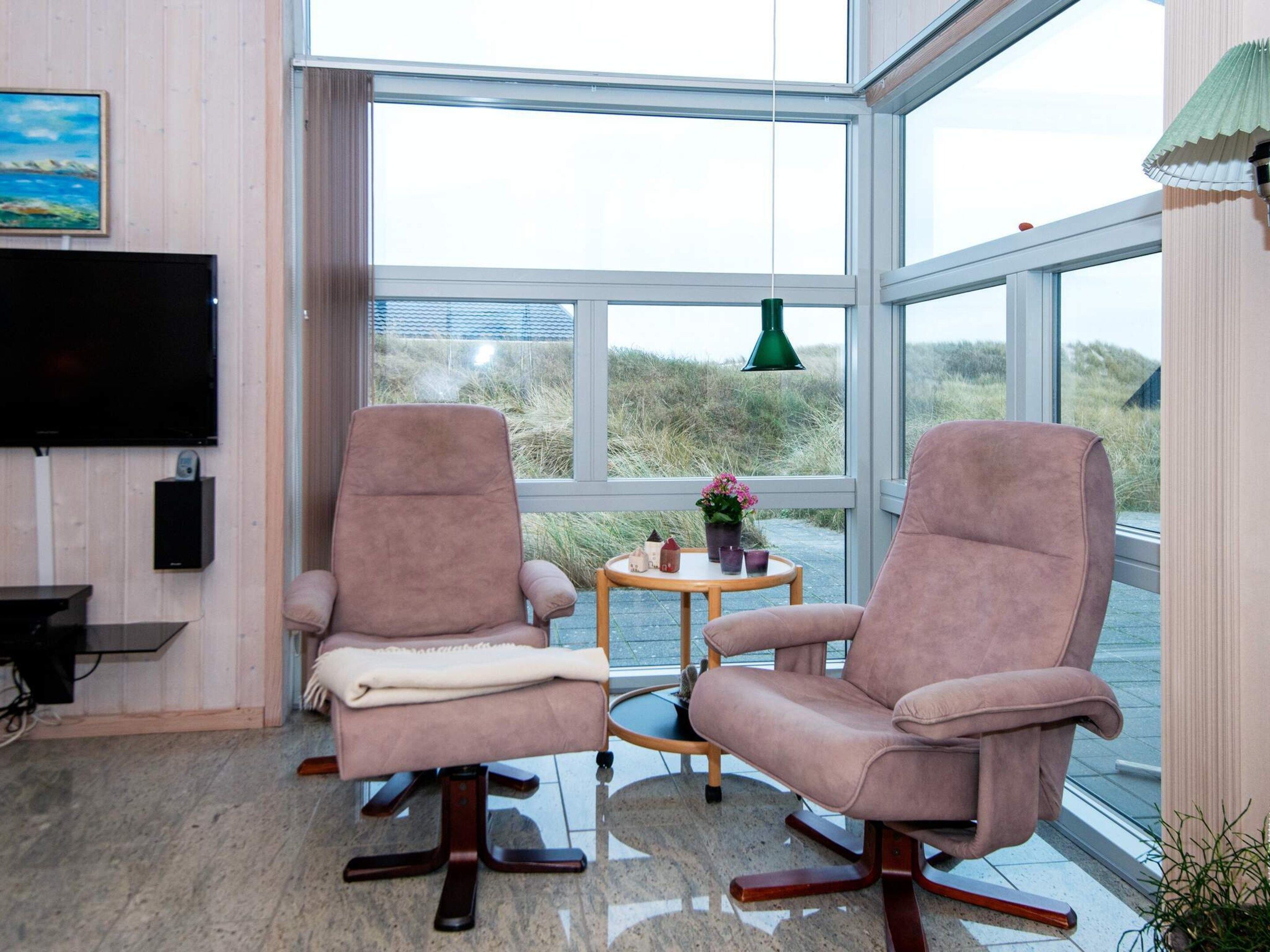A modern room with two cushioned chairs, a small table with a potted plant, a television, and a large window offering a view of the outdoors.