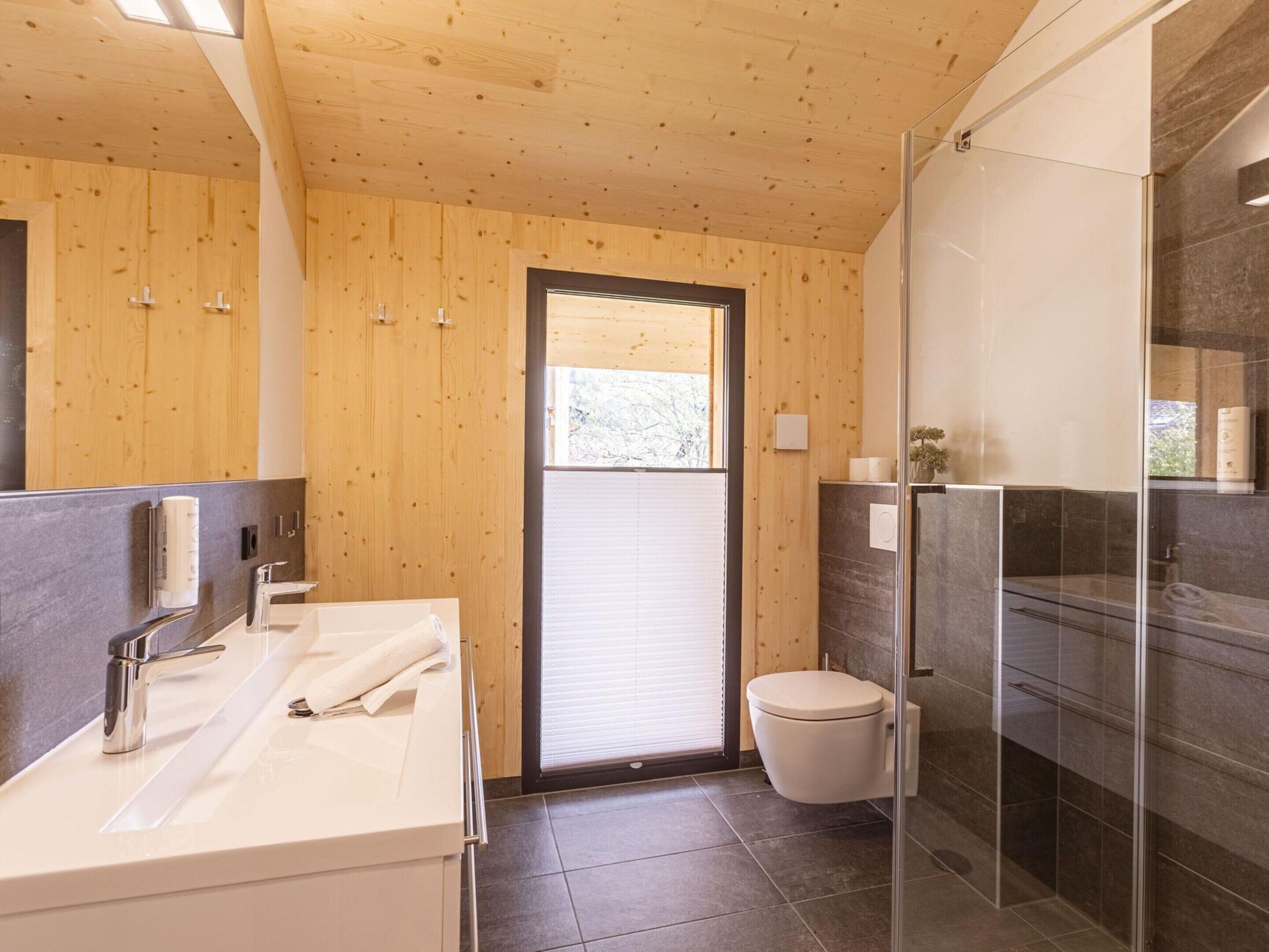 A modern bathroom with wooden walls, a glass shower enclosure, a white sink, and a toilet.