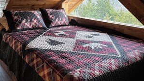 Une chambre avec un plafond en bois, une grande fenêtre et un lit recouvert d’une courtepointe à motifs.
