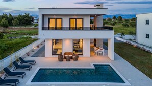 Una casa moderna de dos pisos con piscina, mobiliario exterior y vistas al paisaje circundante.