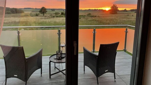A balcony with two wicker chairs and a small table, overlooking a sunset over a field. - Uitzicht op de Kraanvogel (Dreschvitz)