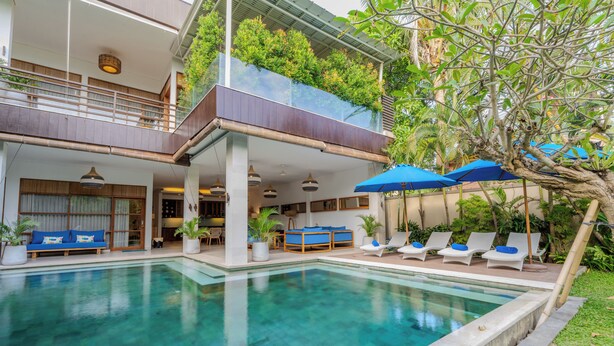 Un área de piscina moderna con sillones de descanso y sombrillas, un espacio de estar techado y un balcón con plantas.
