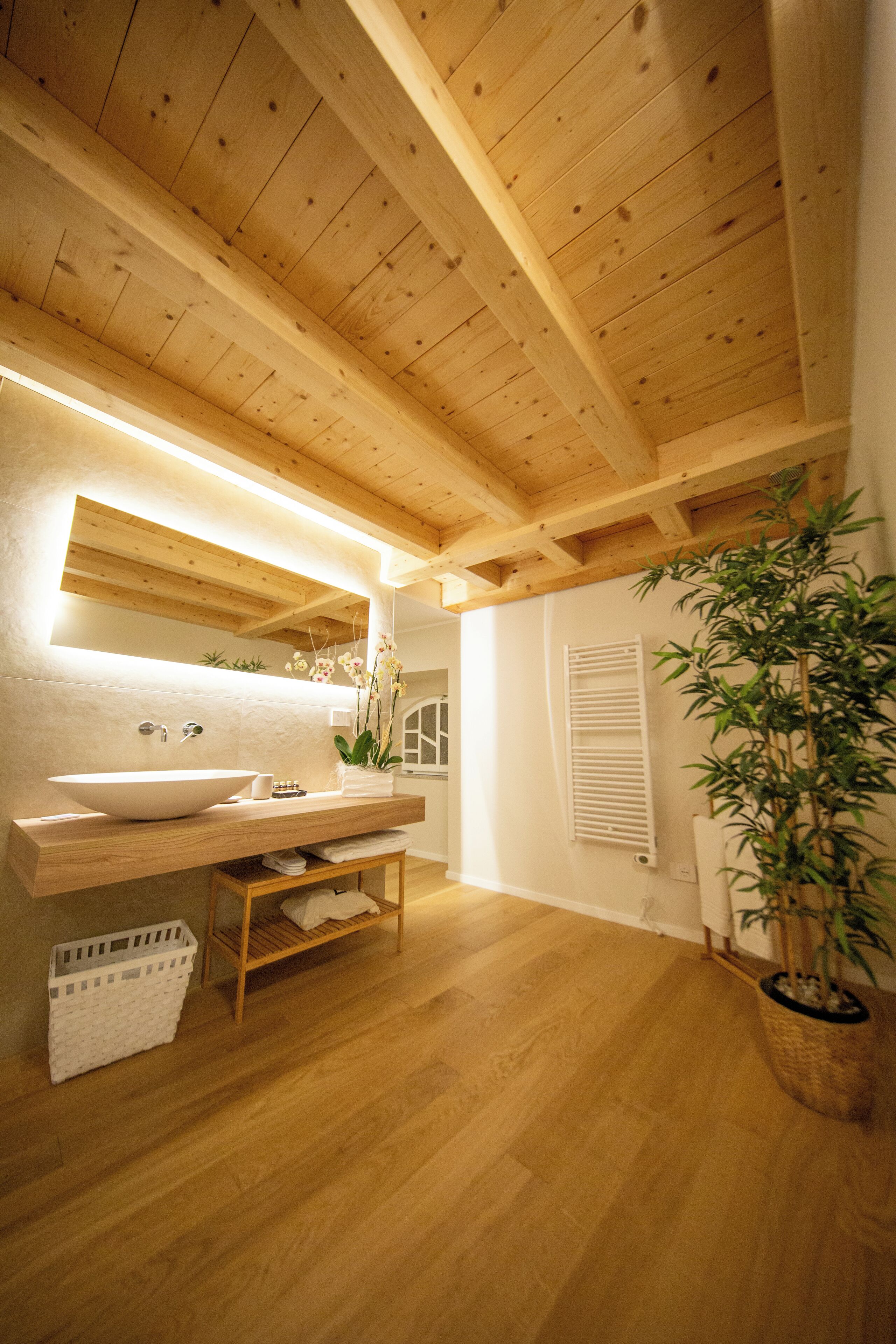 Un plafond en bois, un grand évier blanc, un miroir, une plante et une fenêtre avec des volets.