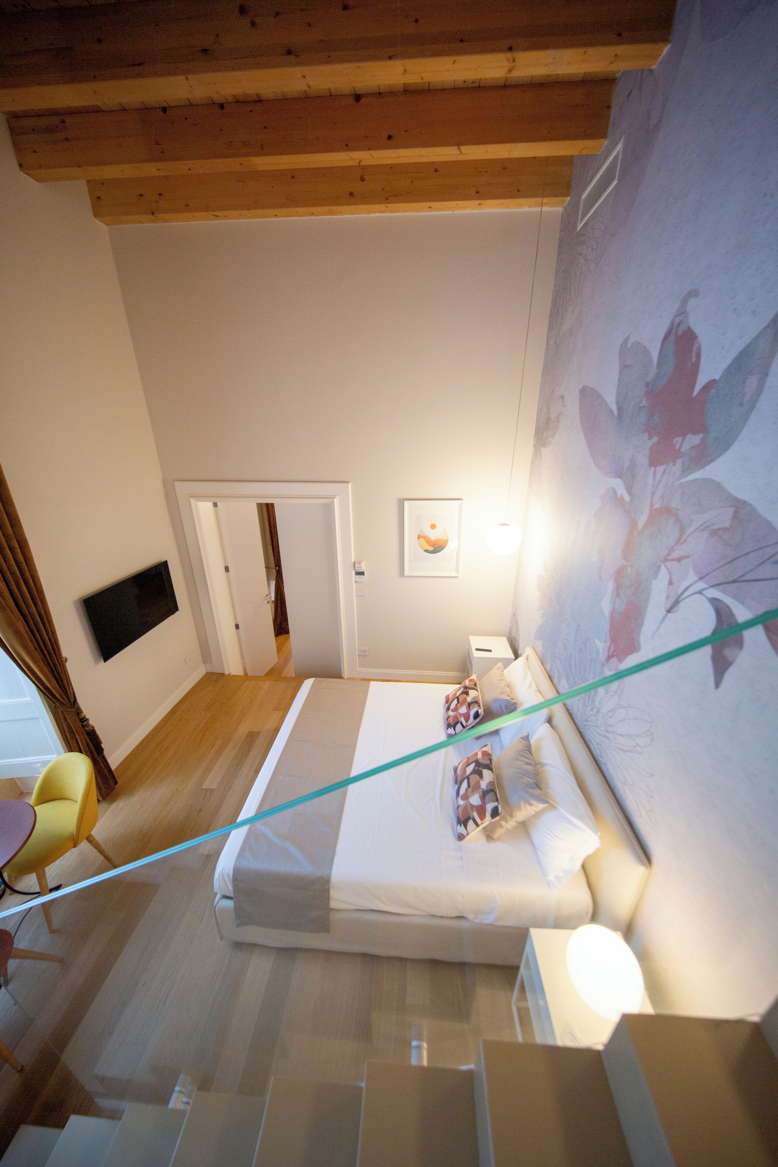 Un dormitorio con cama, televisión montada en la pared, papel tapiz floral y una escalera que conduce a otra habitación.