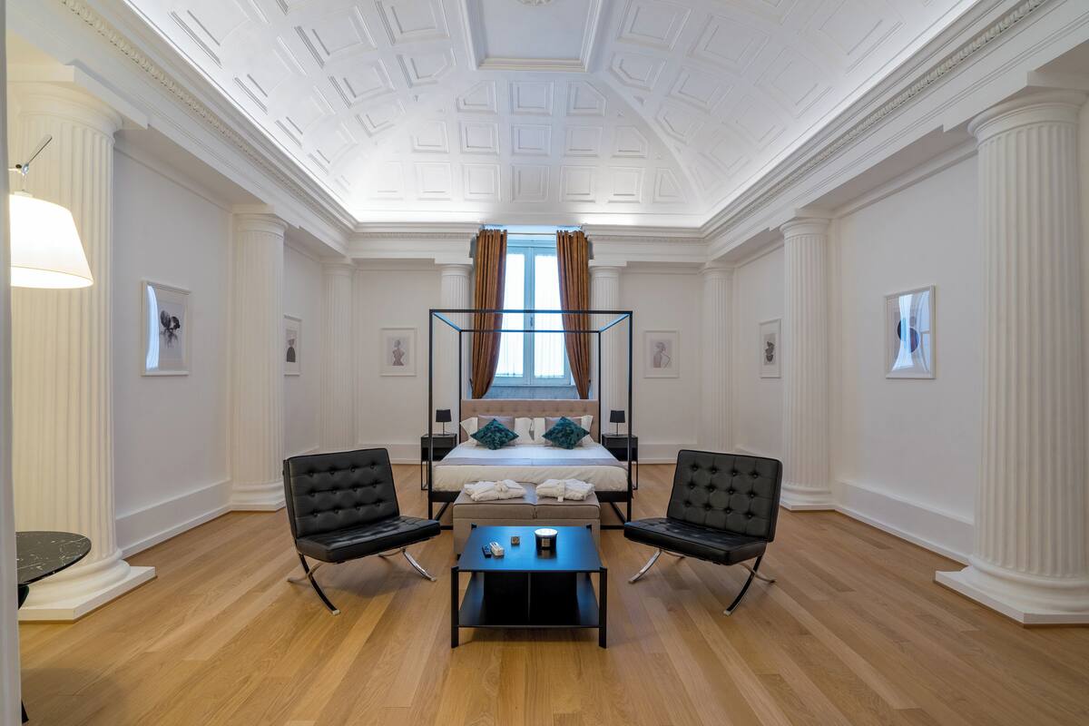 Ein geräumiges Zimmer mit einem großen Bett, zwei Sesseln, einem kleinen Tisch und einer Decke mit dekorativen Stuckverzierungen.