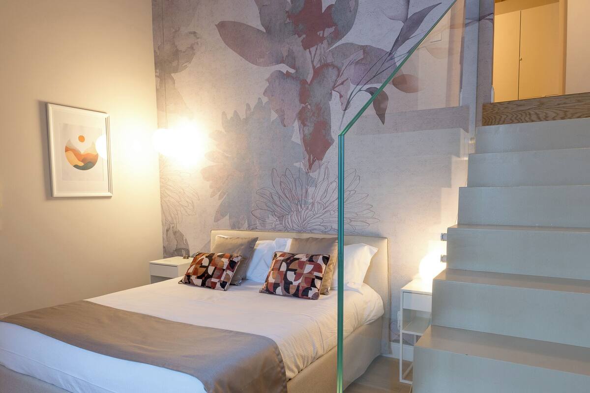 Ein Schlafzimmer mit Bett, Nachttisch, Wandlampe, einer Treppe und einer Wandmalerei mit floralem Dekor.