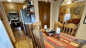 Un comedor con una mesa de madera, sillas y un mantel colorido. Se observa una cocina al fondo, con microondas, horno y refrigerador. La habitación tiene pisos de madera y una lámpara de araña.