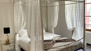 A four-poster canopy bed with white bedding, a bedside table with a lamp, and a window with curtains. - Hotel Restaurant L'Esperance (Saint-Cast-le-Guildo)