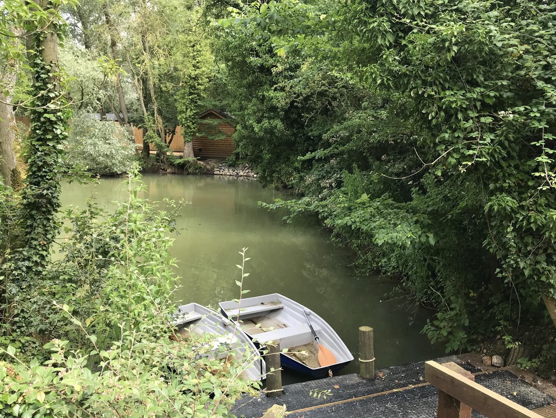 A calm river with boats docked, surrounded by dense greenery and a wooden structure in the background.