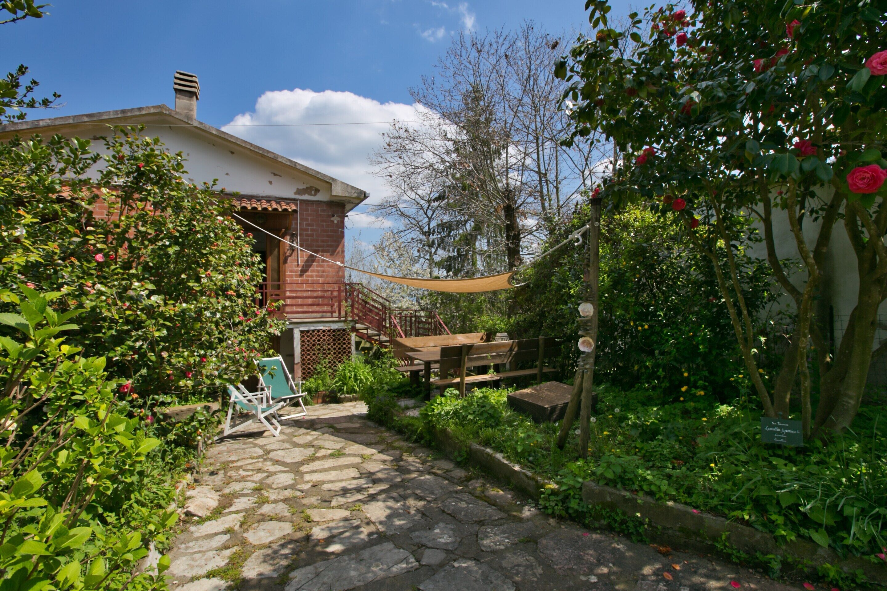 A stone pathway leading to a house with a red roof and a garden with various plants and flowers.