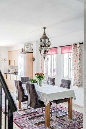 Uma sala de jantar com uma mesa posta para quatro pessoas, rodeada por cadeiras, um candelabro e vista para a cozinha ao fundo.