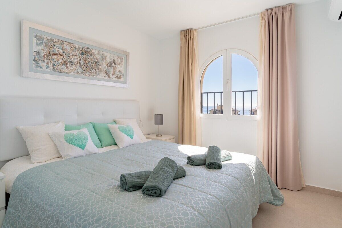 A bedroom with a bed, pillows, a nightstand, a lamp, a window with curtains, and a painting on the wall.
