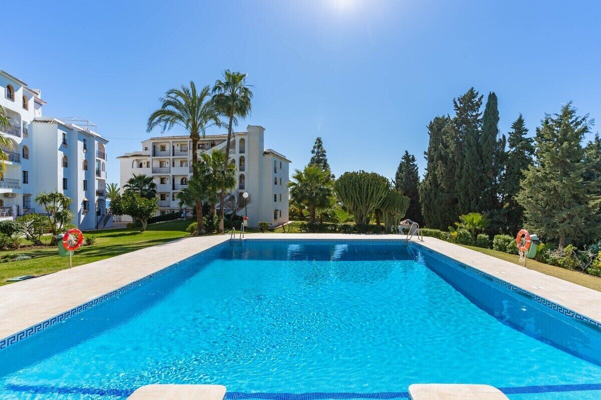 A rectangular swimming pool with clear blue water, surrounded by a paved deck. There are palm trees and other greenery in the background, and a building with multiple balconies is visible to the left.