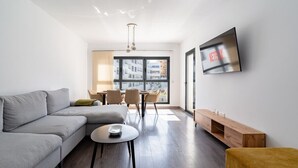 Een moderne woonkamer met een grijze bank, een klein rond tafeltje en een aan de muur bevestigde televisie.