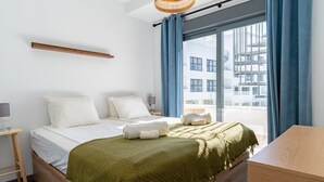 Een moderne slaapkamer met een groot bed, een houten hoofdeinde, een nachtkastje met een lamp en uitzicht op een gebouw door een raam met blauwe gordijnen.